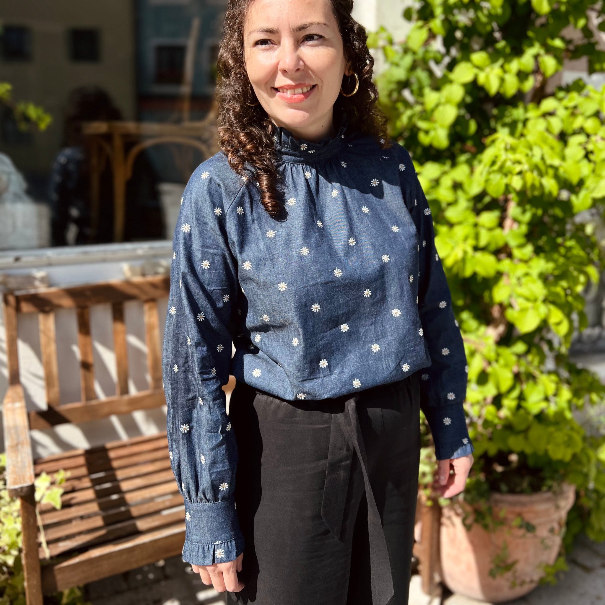 Frau in blauer Bluse mit Blumenmuster und schwarzem Rock steht draußen vor einer Bank, umgeben von grünen Pflanzen.