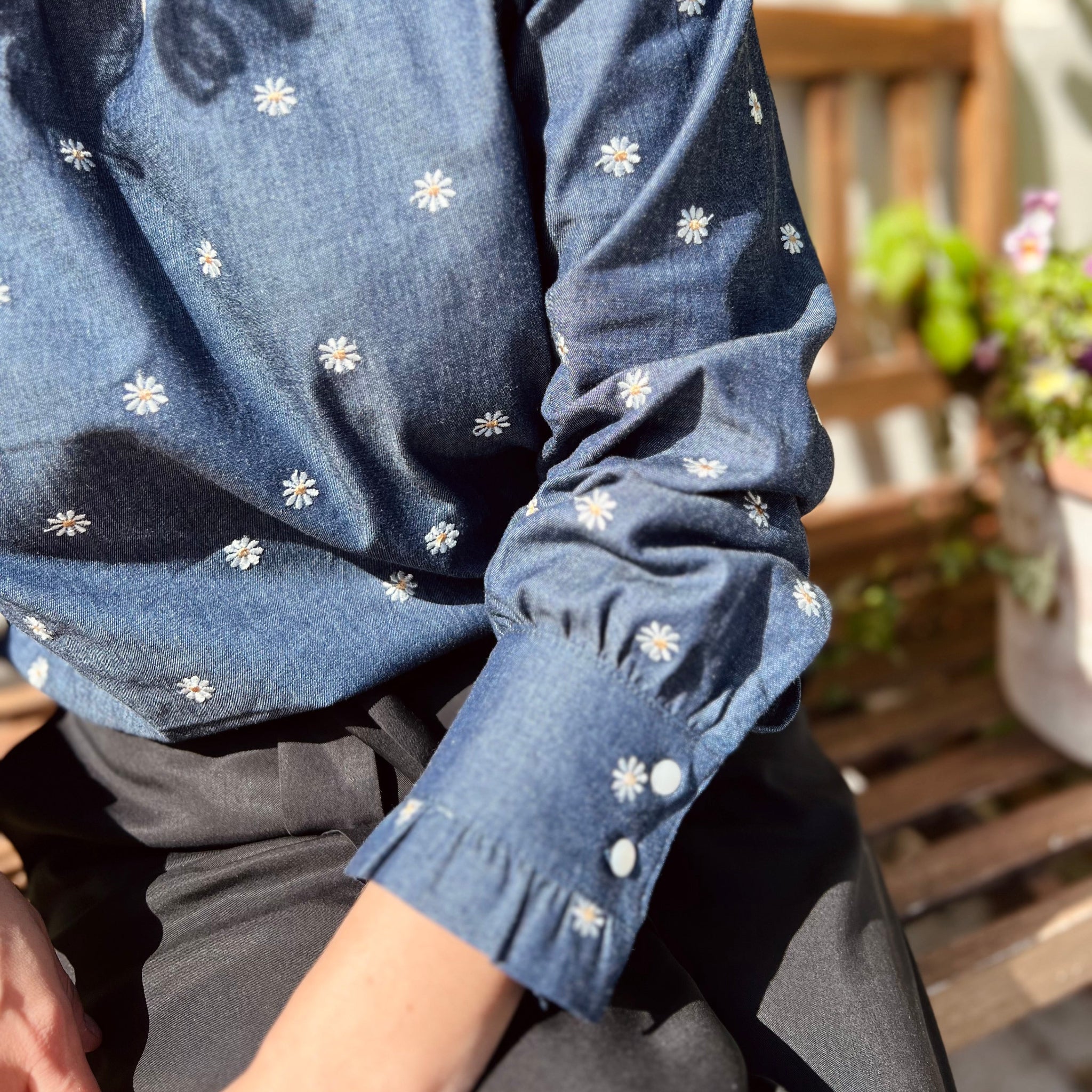 Person trägt eine blaue Bluse mit weißen Blumendetails, sitzt auf einer Holzbank neben einer Pflanze.