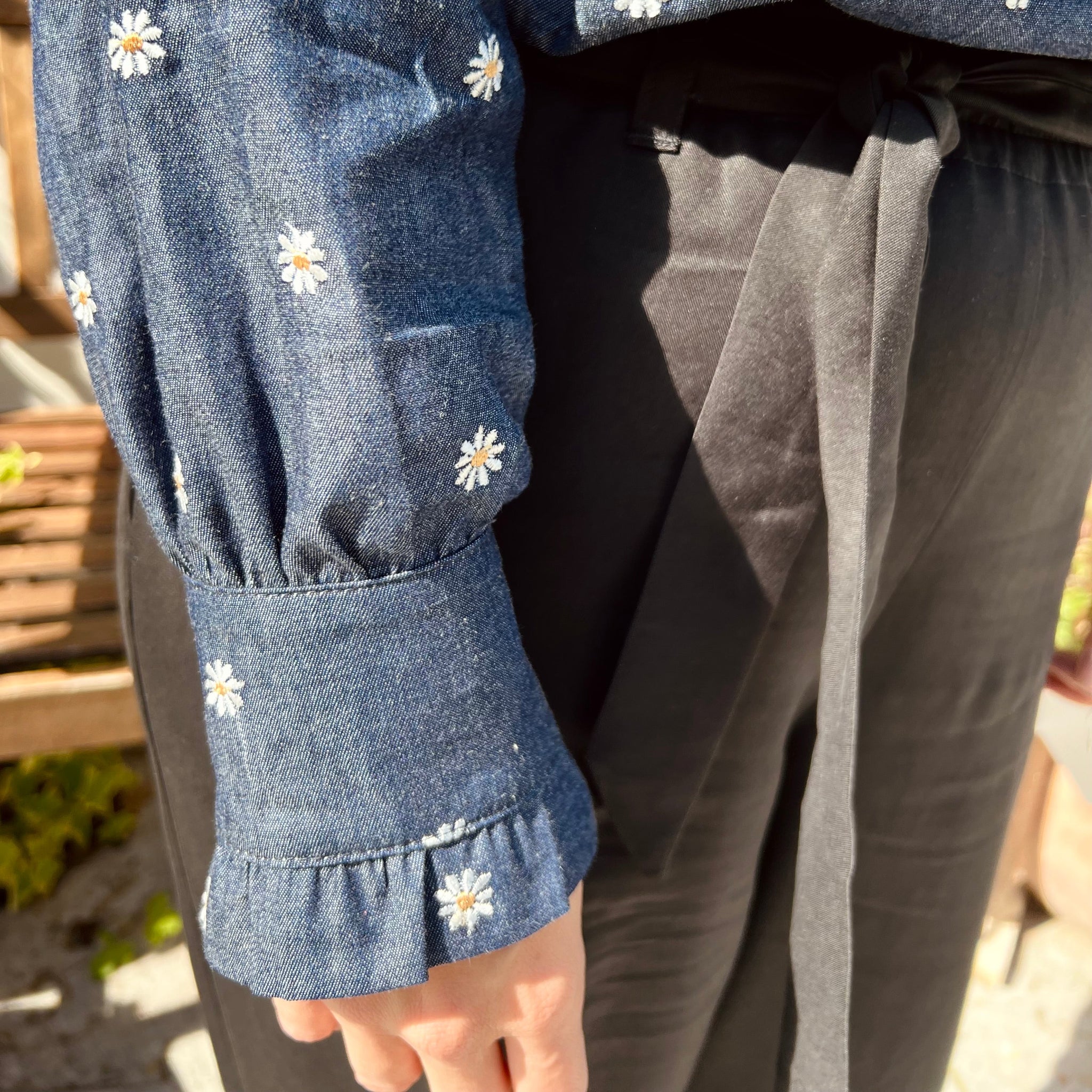 Detail einer Person im Freien, die ein dunkelblaues Hemd mit Gänseblümchenstickerei und schwarze Hose trägt.