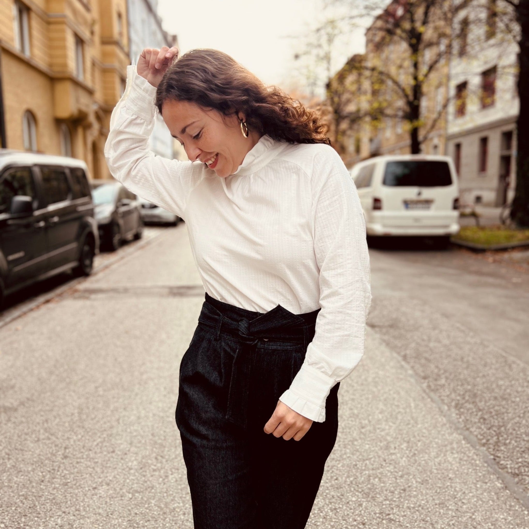 Eine Frau mit weißer Bluse und schwarzen Hosen lächelt und geht fröhlich in einer städtischen Straße.