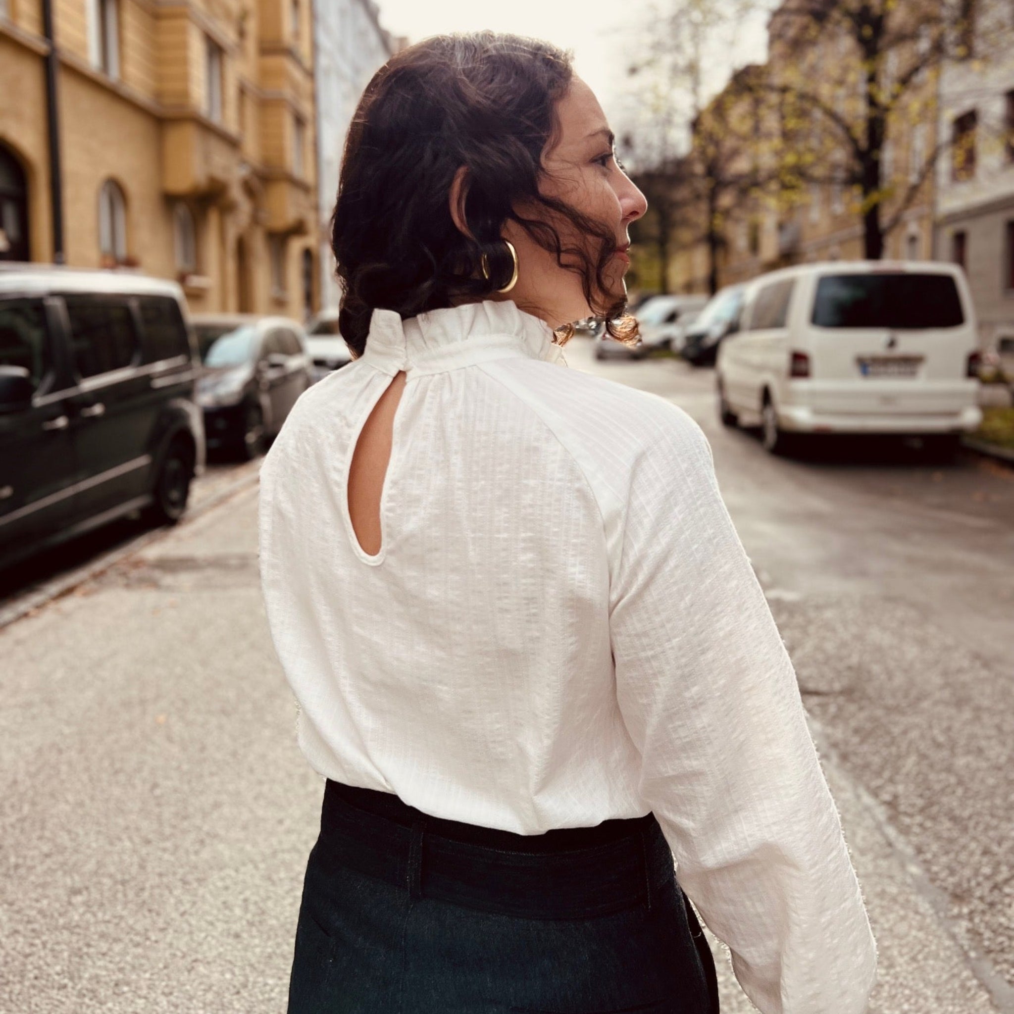 Frau mit lockigem Haar steht auf einer städtischen Straße, trägt eine weiße Bluse mit Rückenausschnitt.
