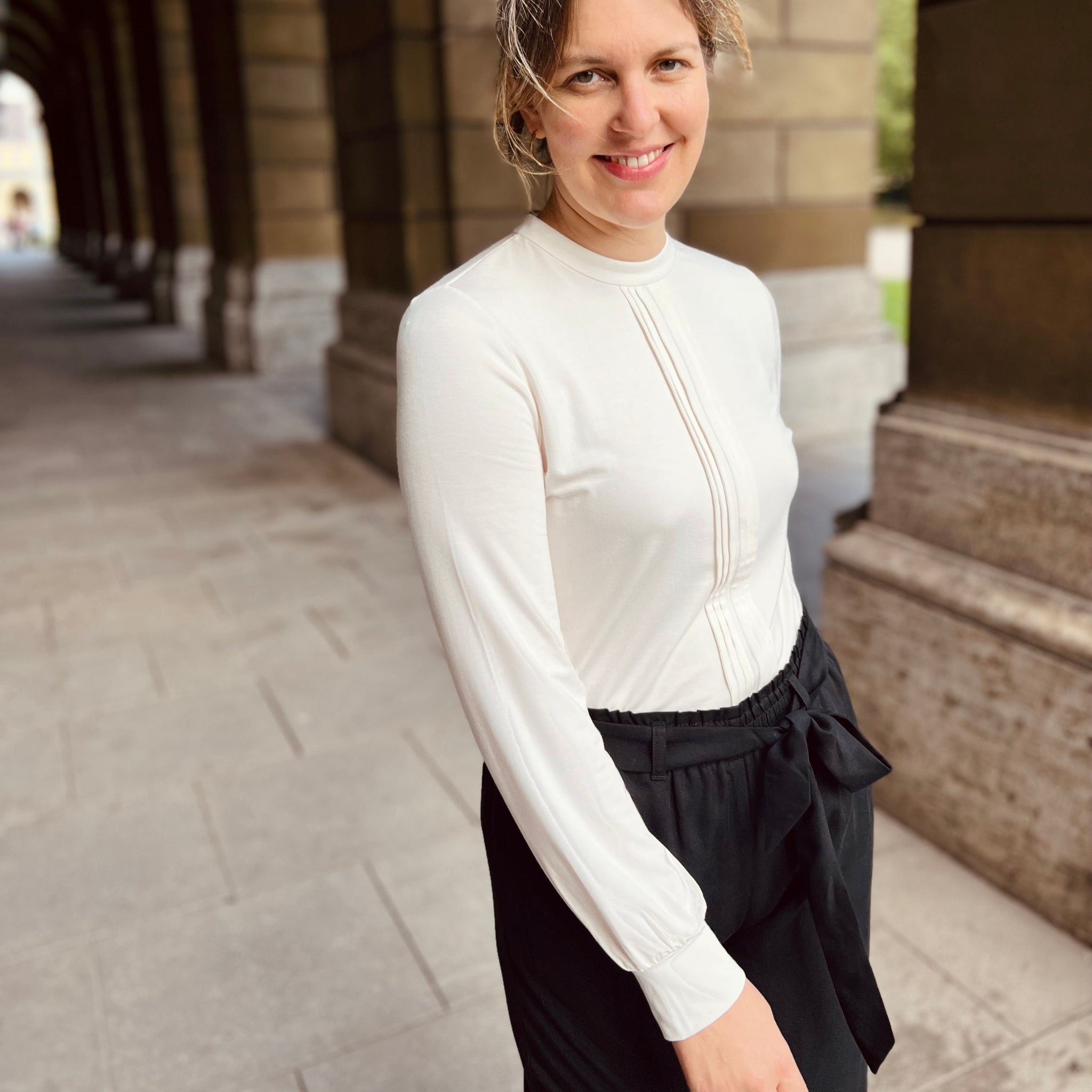 Eine Frau in einem weißen Oberteil und schwarzer Hose lächelt in einem Steingang mit gewölbten Pfeilern.