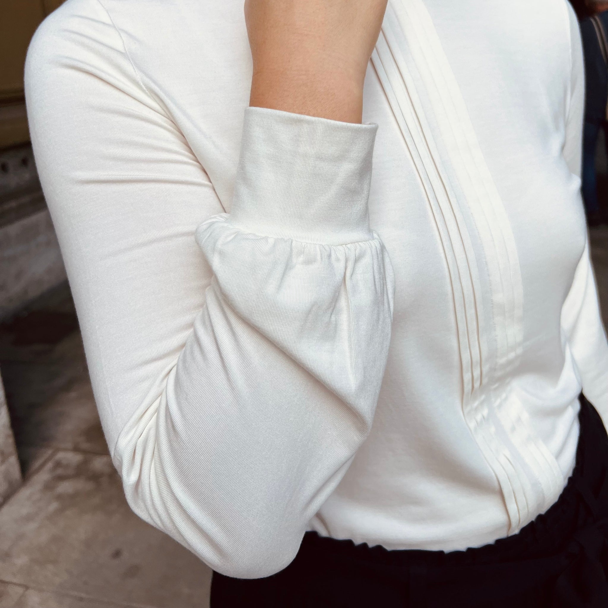 Person in a white long-sleeve blouse with pleated details, hand raised towards their face.