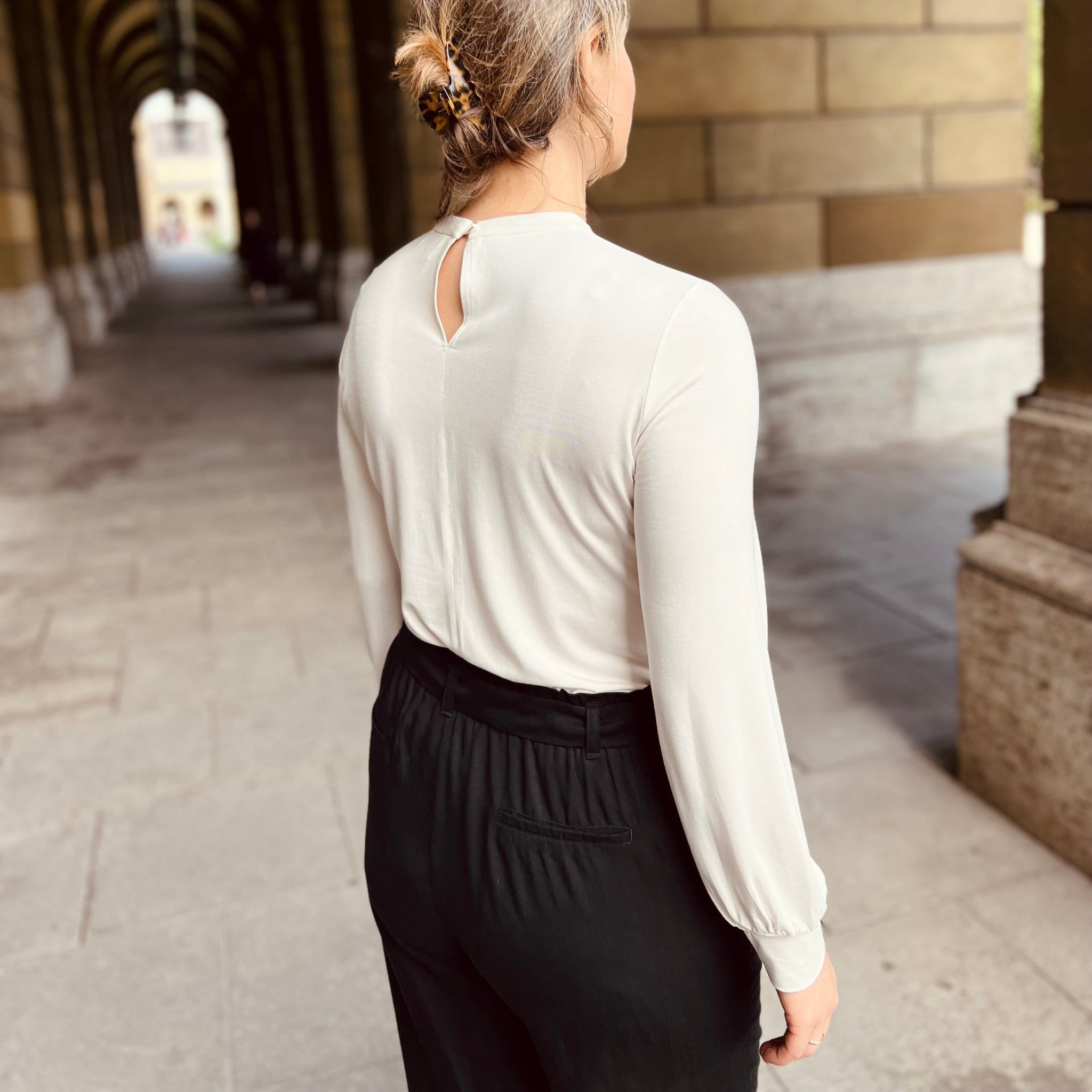 Eine Frau mit einem Dutt geht durch einen Arkadengang, trägt eine weiße Bluse und schwarze Hose.