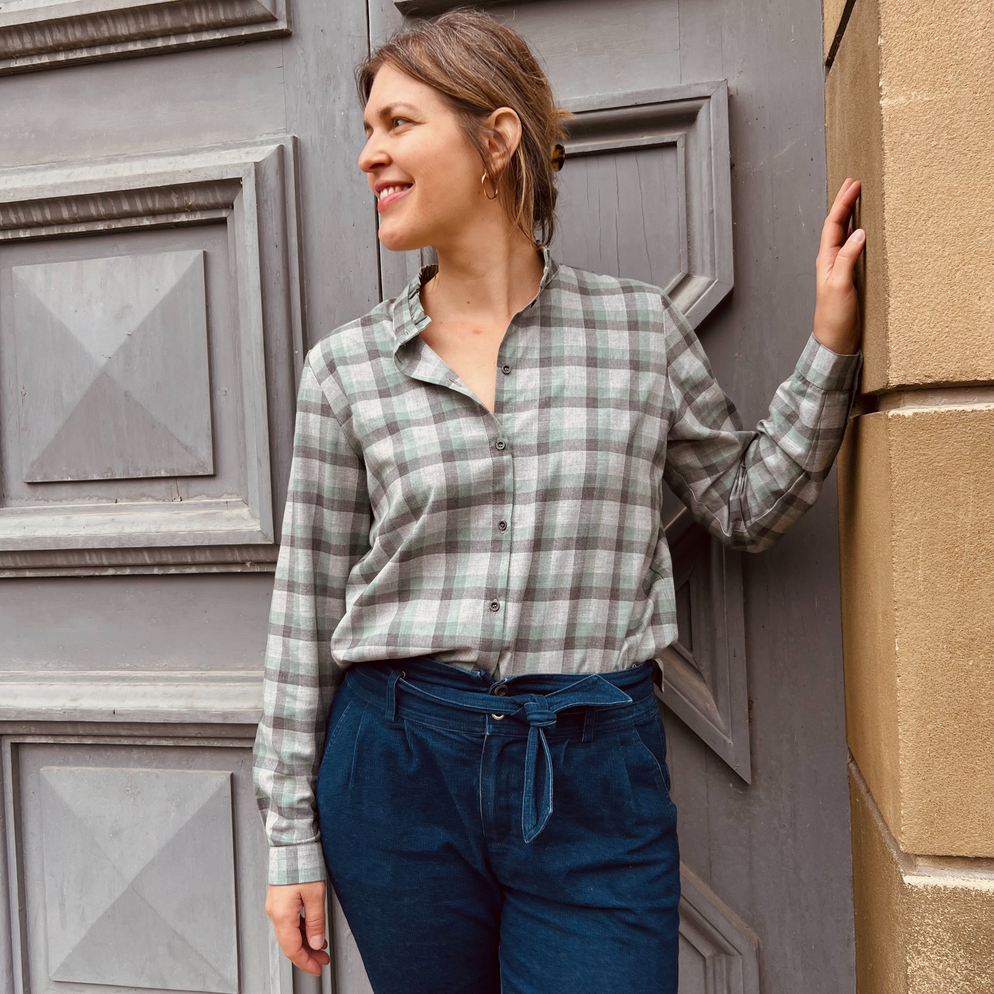 Frau in kariertem Hemd und blauer Hose lehnt lächelnd an einer Holztür und blickt zur Seite.