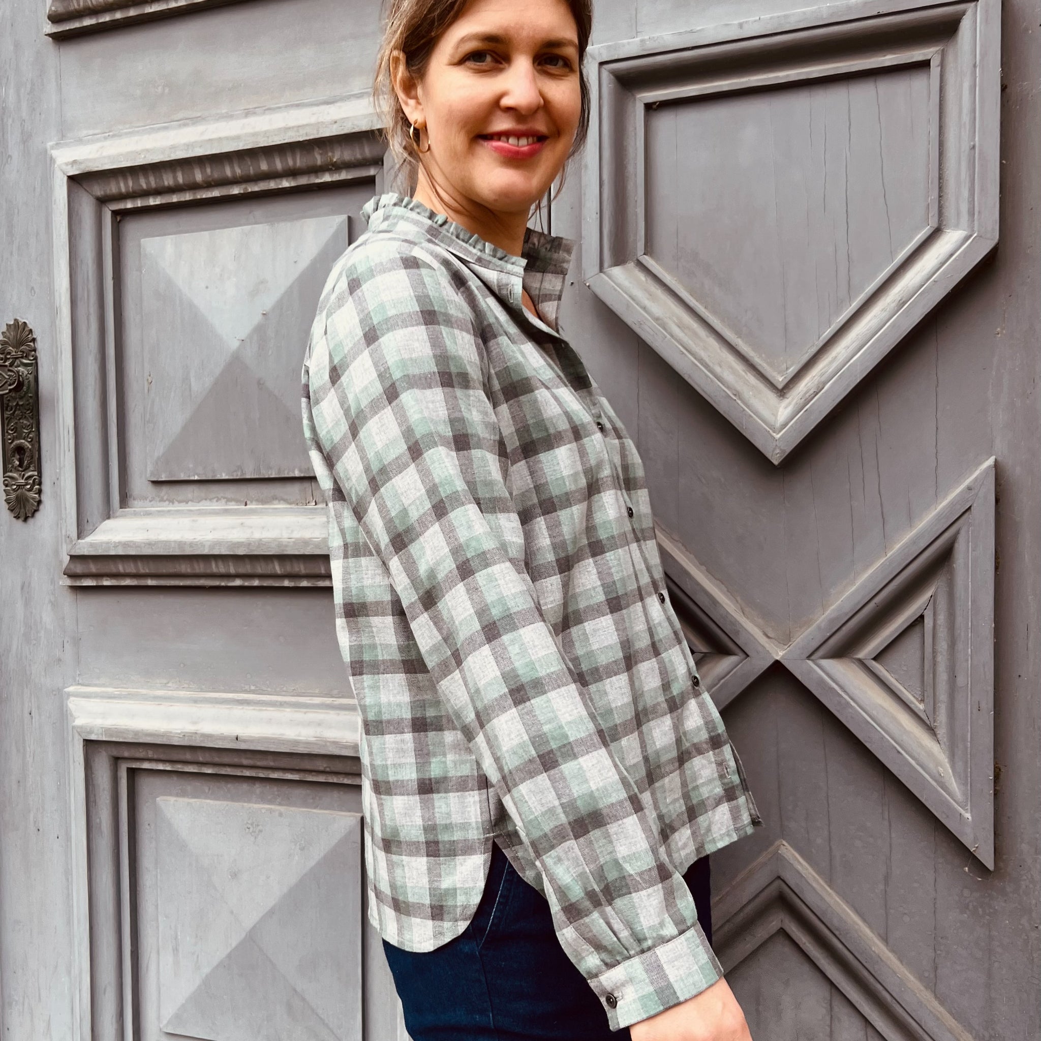 Frau in kariertem Hemd steht vor einer detaillierten Holztür und lächelt in die Kamera.