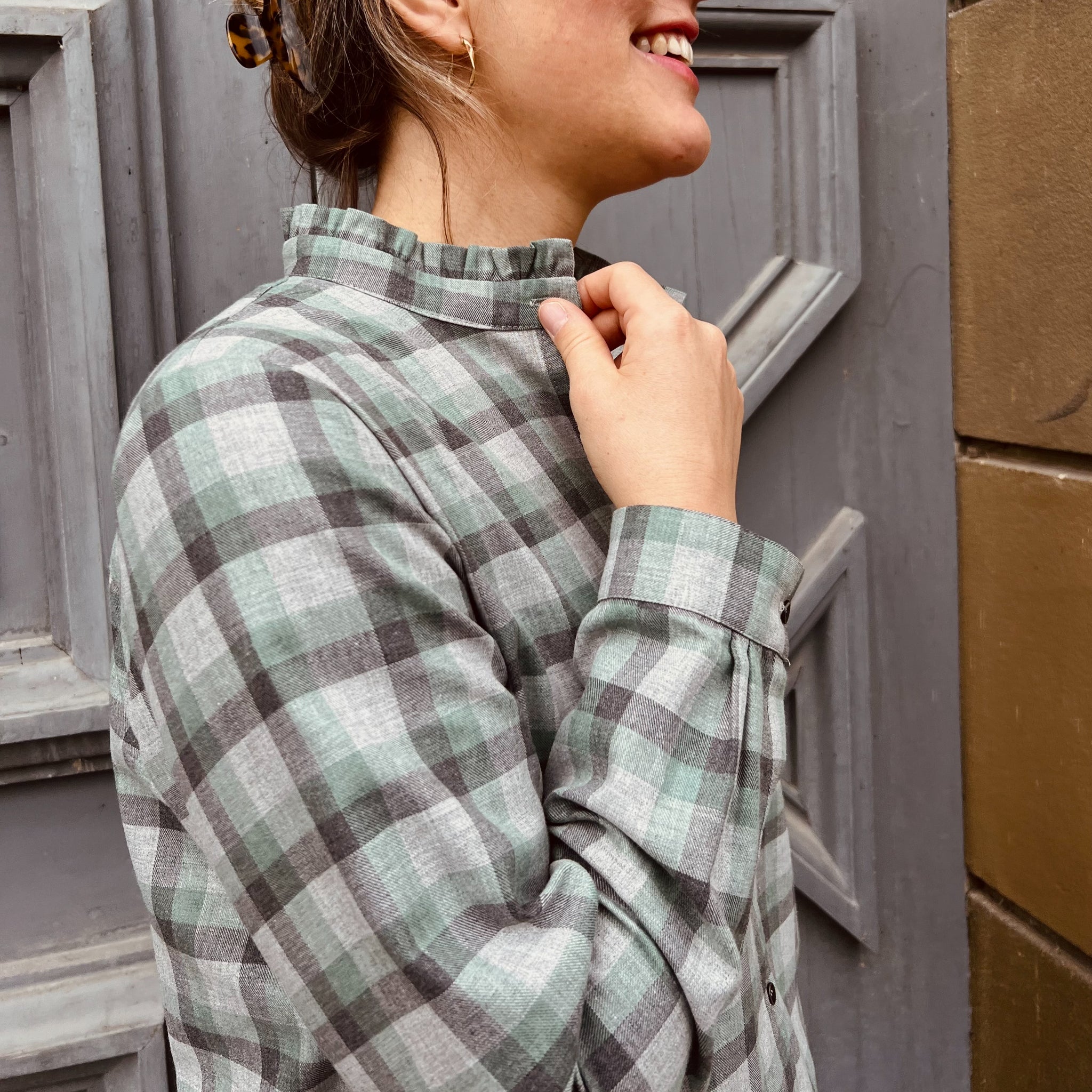 Frau in kariertem Hemd steht lächelnd vor einer grauen Tür und hebt den Kragen mit der Hand an.