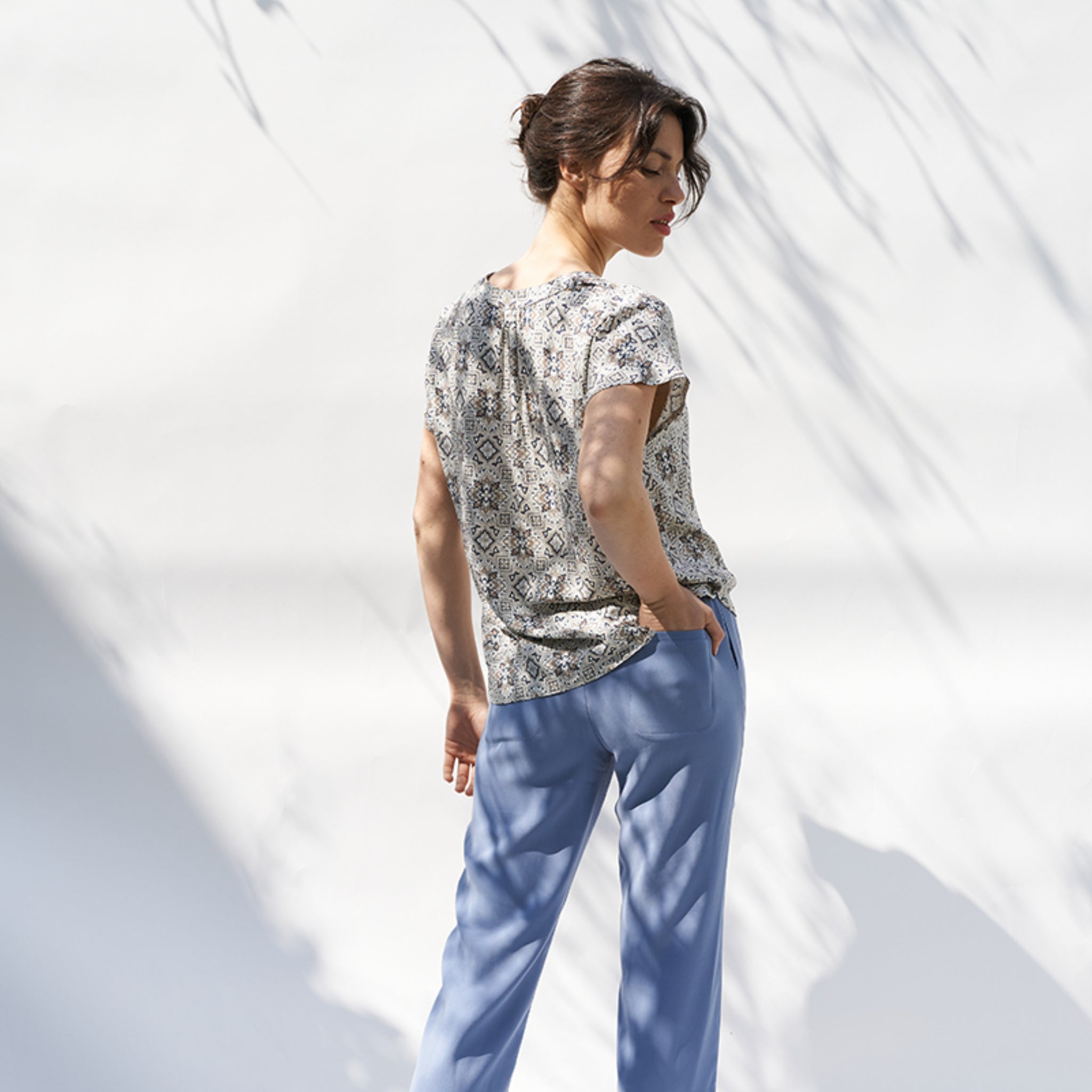 Eine Frau steht seitlich in blauer Hose und gemustertem Oberteil vor einer weißen Wand mit Schatten von Zweigen.