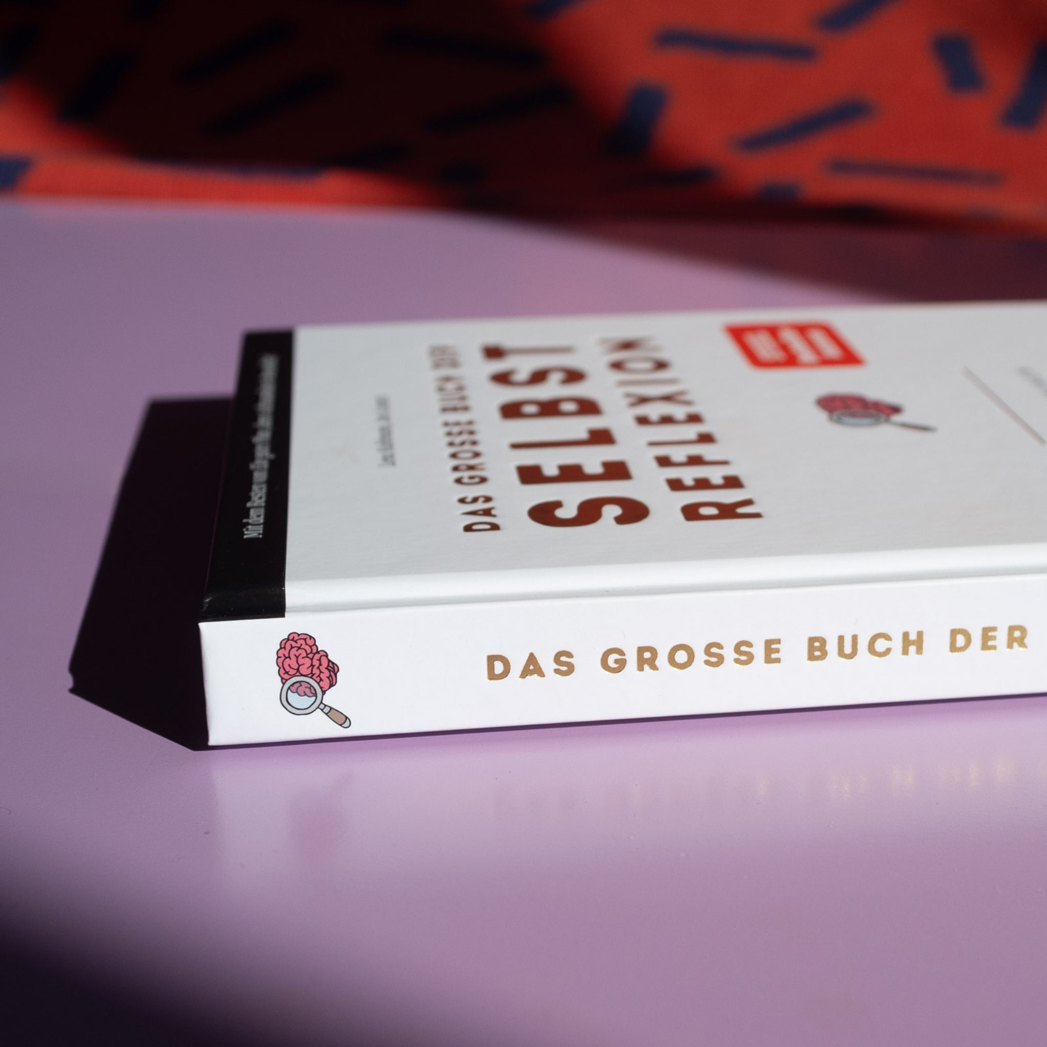 Nahaufnahme eines Buches mit dem Titel "Das große Buch der Selbstreflexion" auf einem lila Tisch.