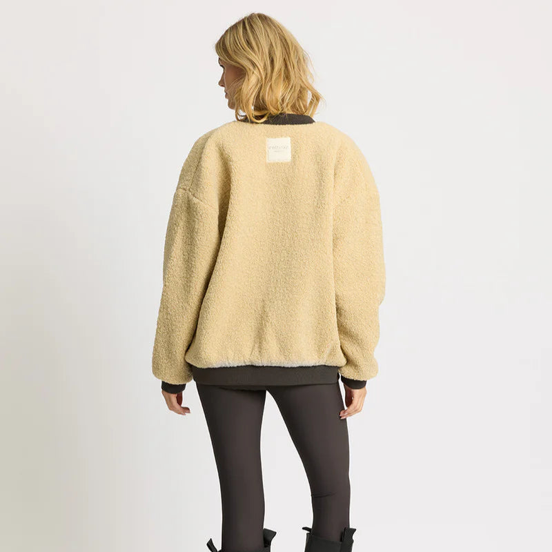 Eine Person mit schulterlangem blondem Haar steht mit dem Gesicht zur Wand, trägt eine lockere beigefarbene Fleecejacke mit einem Aufnäher auf dem oberen Rücken und schwarze Leggings und steht vor einem schlichten hellen Hintergrund.