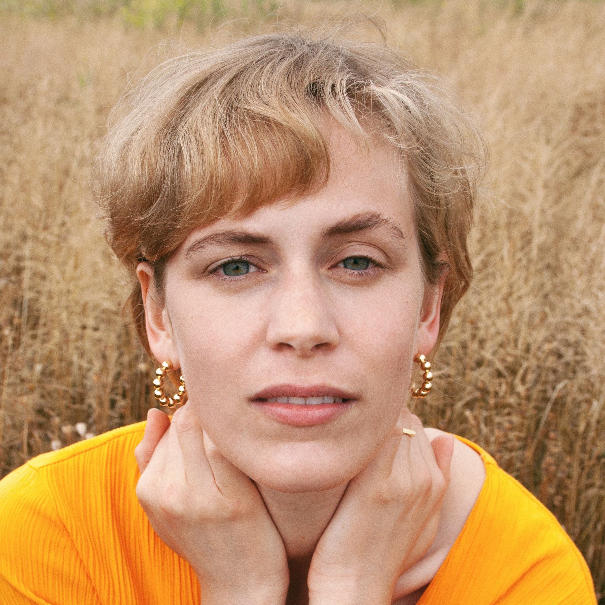 Eine Person mit kurzen blonden Haaren, die in einem goldenen Shirt auf einem Feld posiert, stützt ihr Gesicht in die Hände.