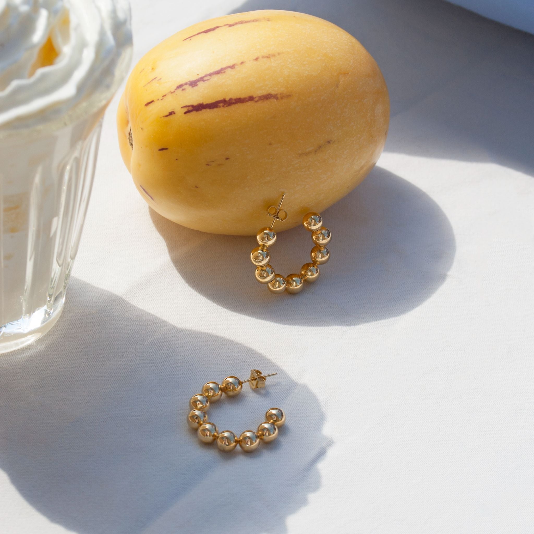 Goldene Creolen liegen neben einer gelben Frucht auf einem weißen Tisch, daneben ein Glas mit Sahne.