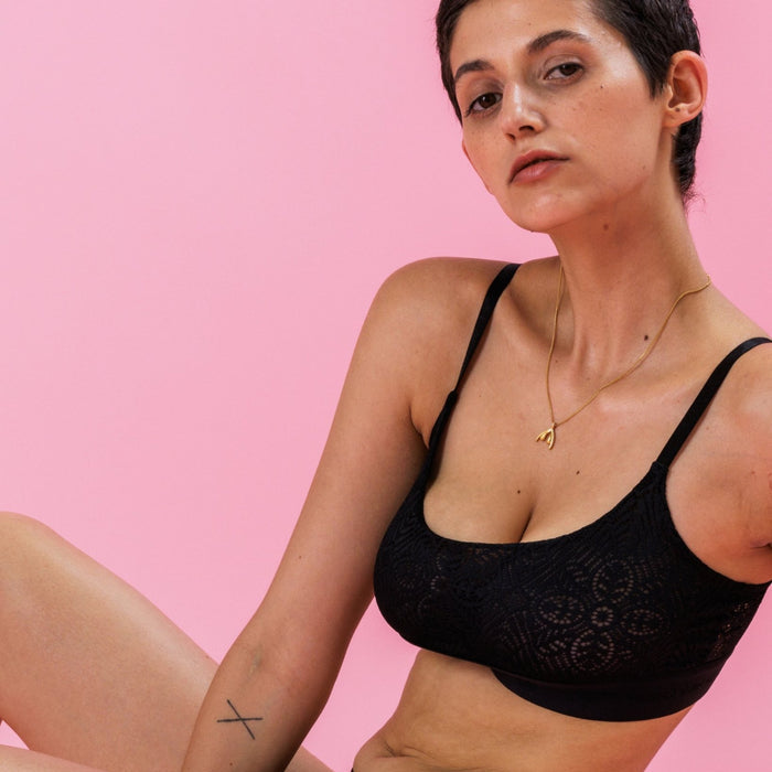 Frau in schwarzen Spitzendessous vor rosa Hintergrund, mit kurzem Haar und feinem Goldschmuck.