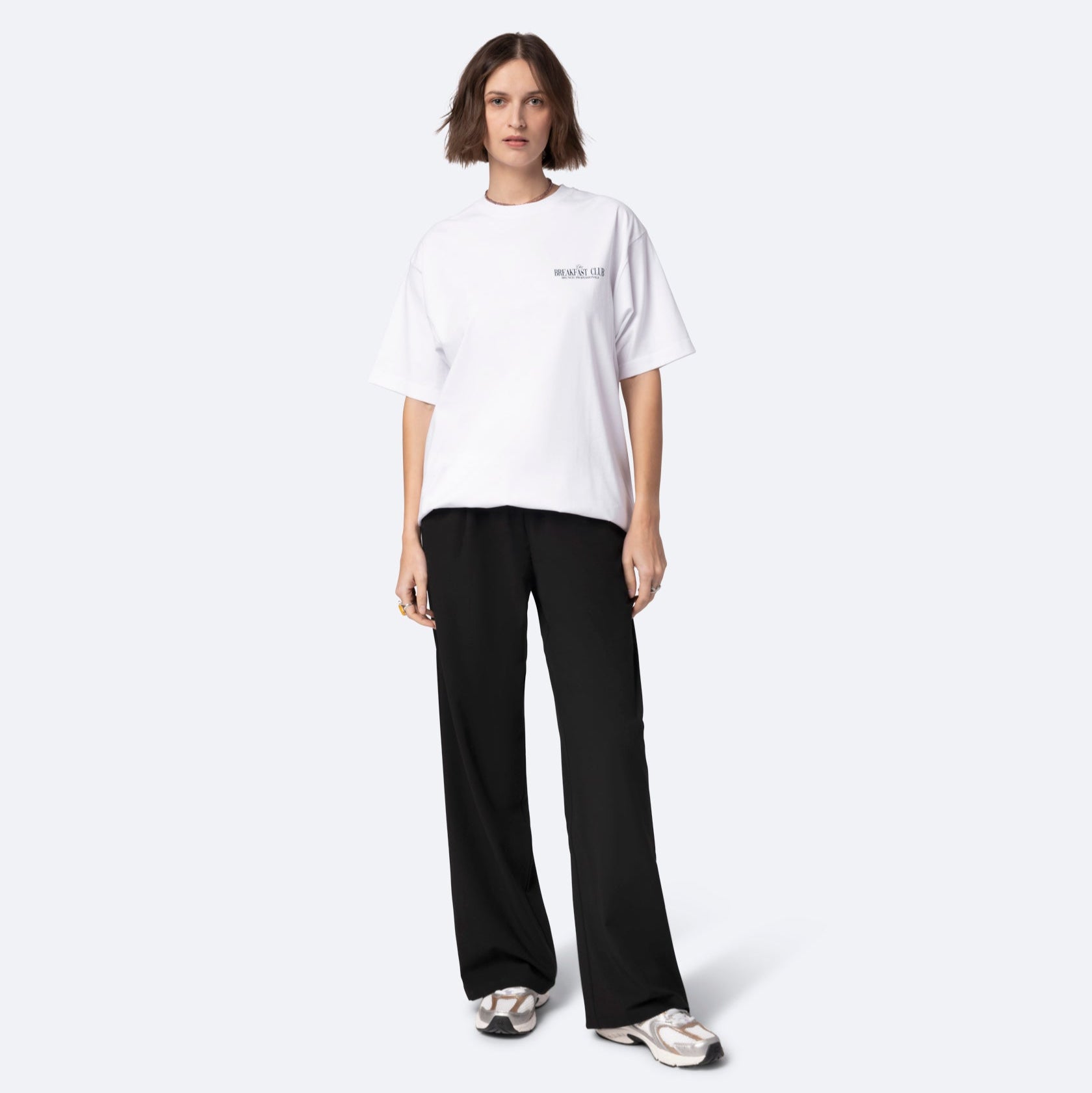 Eine Frau mit kurzen braunen Haaren trägt ein lockeres weißes T-Shirt, eine schwarze Hose mit weitem Bein und weiße Turnschuhe. Sie steht vor einem schlichten hellen Hintergrund und blickt mit neutralem Gesichtsausdruck nach vorne.