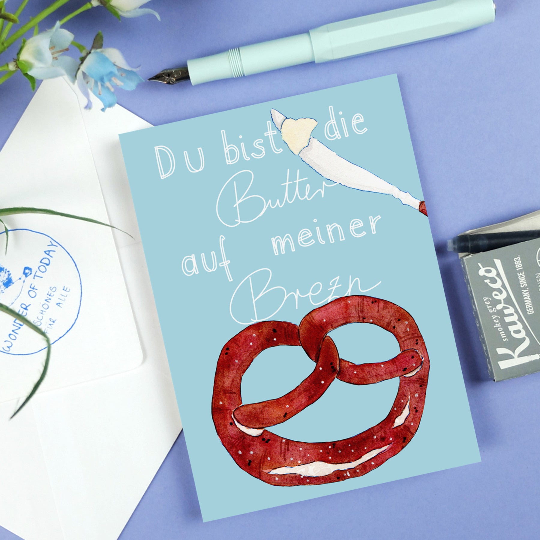 Eine Grußkarte auf blauem Hintergrund mit einer Illustration einer Butterbrezel und dem deutschen Text Du bist die Butter auf meiner Brezn. In der Nähe befinden sich ein Stift, ein Umschlag und ein Stempelkissen.