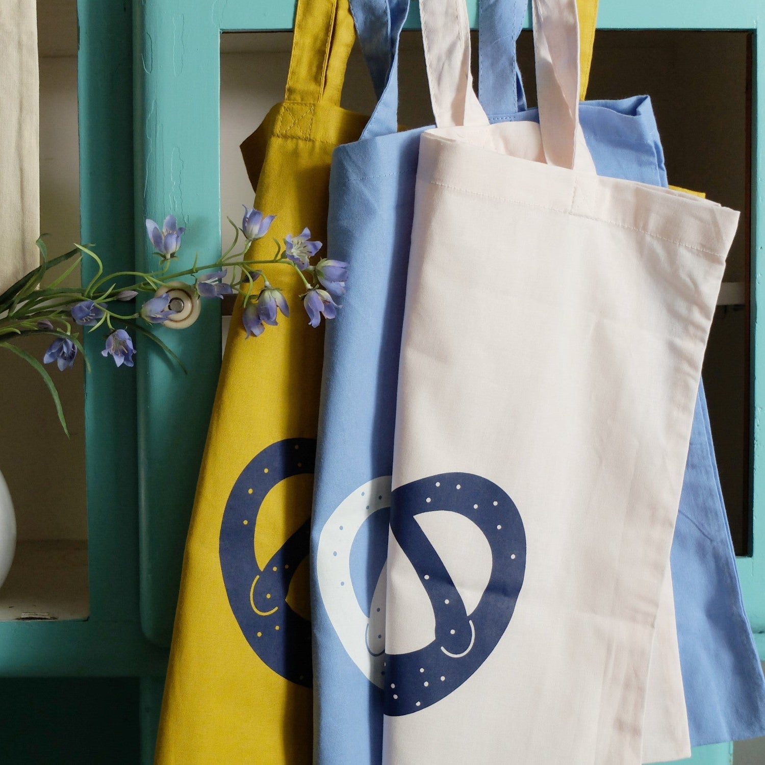 Drei Tragetaschen aus Stoff - gelb, blau und grauweiß - hängen nebeneinander. Die gelben und blauen Taschen sind mit einem Brezelmuster versehen. Hinter den Taschen befindet sich ein lilafarbener Blumenzweig auf türkisfarbenem Hintergrund.