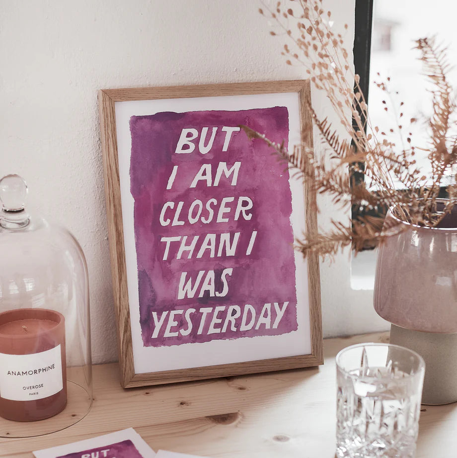 Ein Holzrahmen mit lila Text "BUT I AM CLOSER THAN I WAS YESTERDAY" steht auf einem Tisch mit Kerze und Glas.