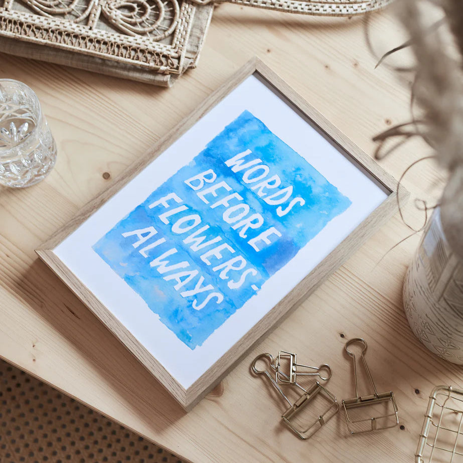 Gerahmtes Bild mit blauer Schrift auf einem Holztisch; daneben ein Glas, Ordnerklammern und dekorative Gegenstände.