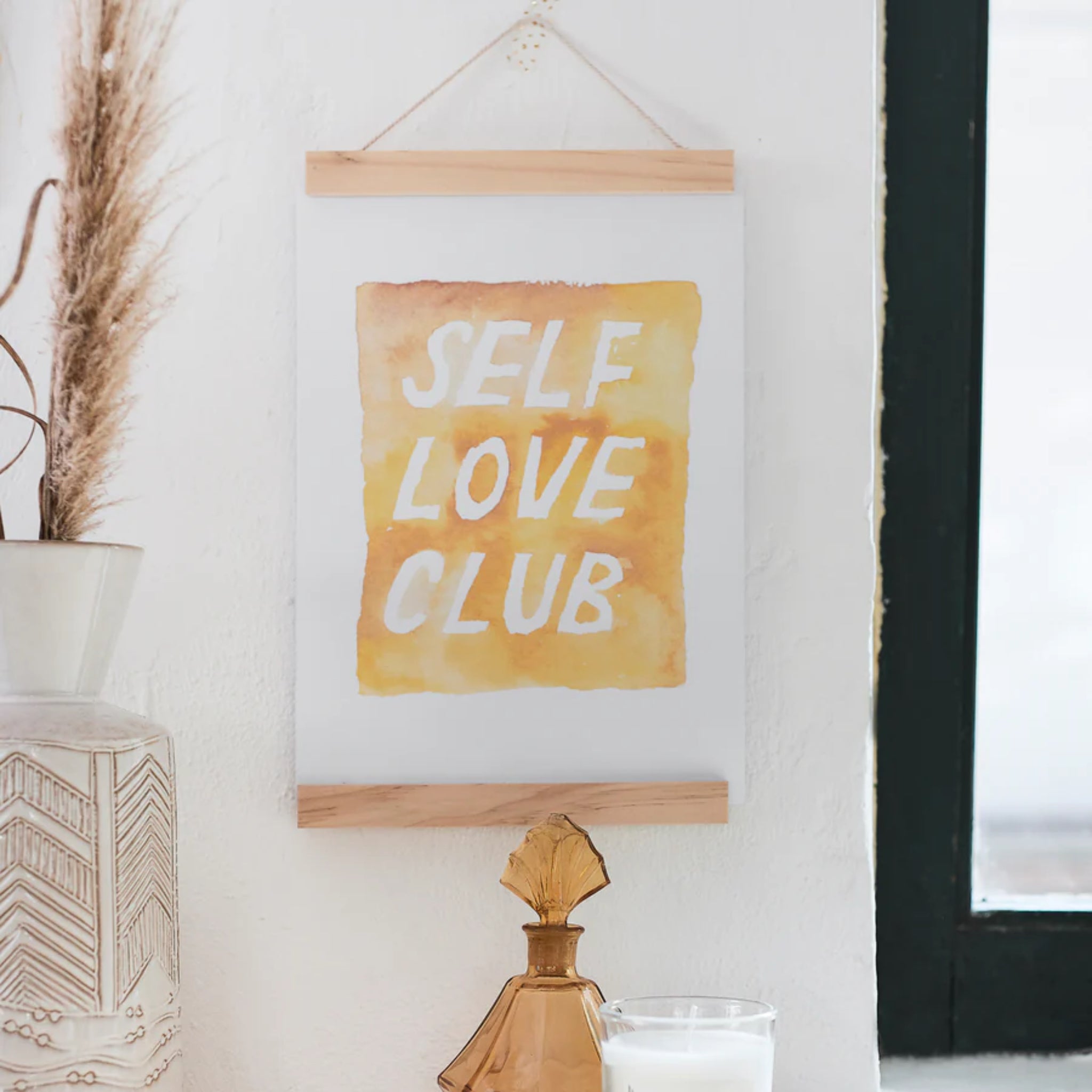 An einer weißen Wand hängt ein Bild mit der Aufschrift "SELF LOVE CLUB" in gelben und orangefarbenen Tönen.