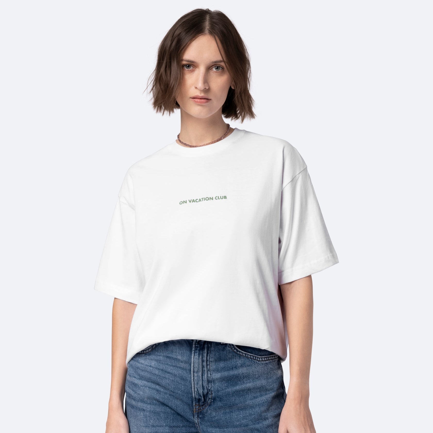 Eine Frau mit kurzen braunen Haaren trägt ein lockeres weißes T-Shirt mit der Aufschrift ON VACATION CLUB und blaue Jeans und steht vor einem schlichten hellen Hintergrund.