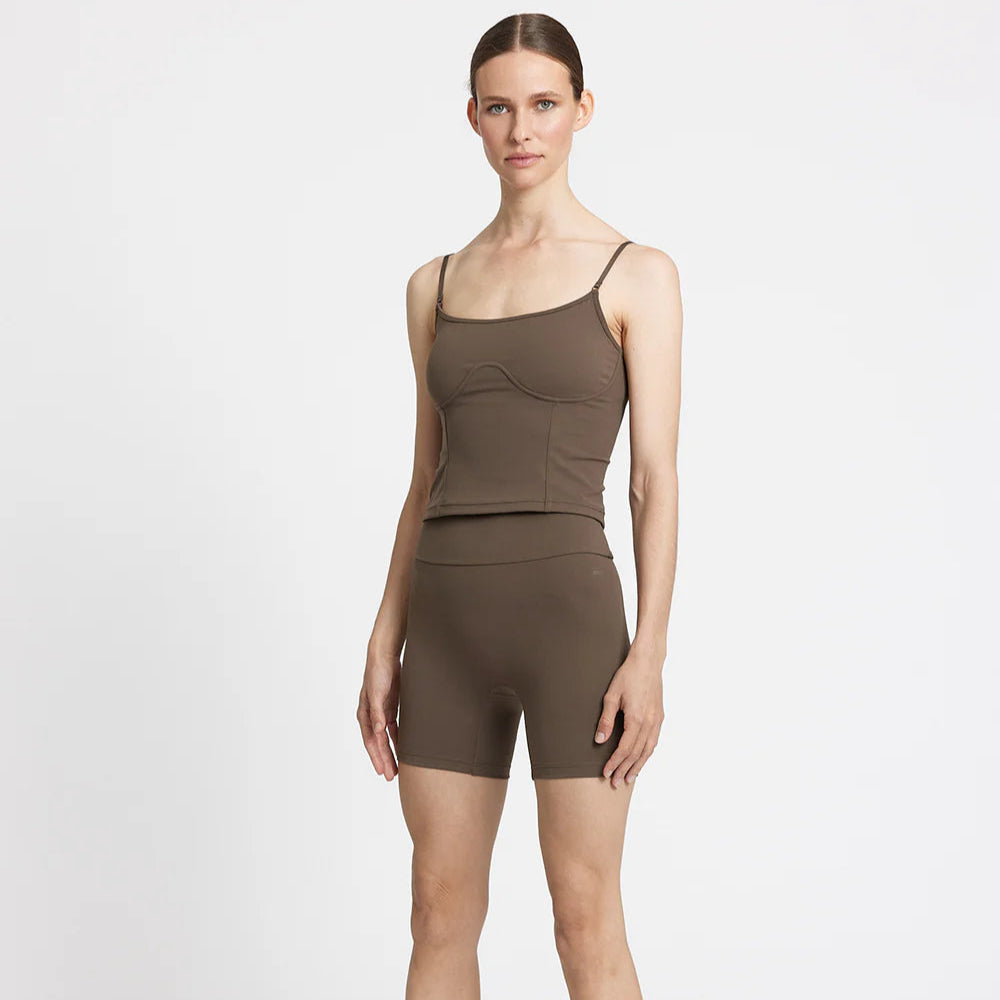 Frau in braunem Fitness-Outfit mit Top und Shorts posiert vor weißem Hintergrund.