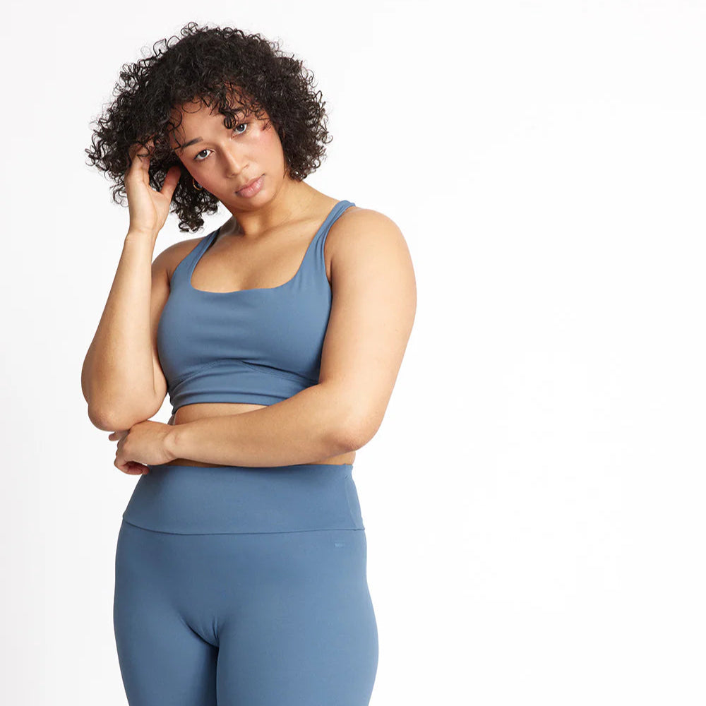 Eine Person mit lockigem Haar, die einen blauen Sport-BH und dazu passende Leggings mit hoher Taille trägt, steht vor einem schlichten weißen Hintergrund und blickt in die Kamera, während eine Hand ihr Haar berührt.