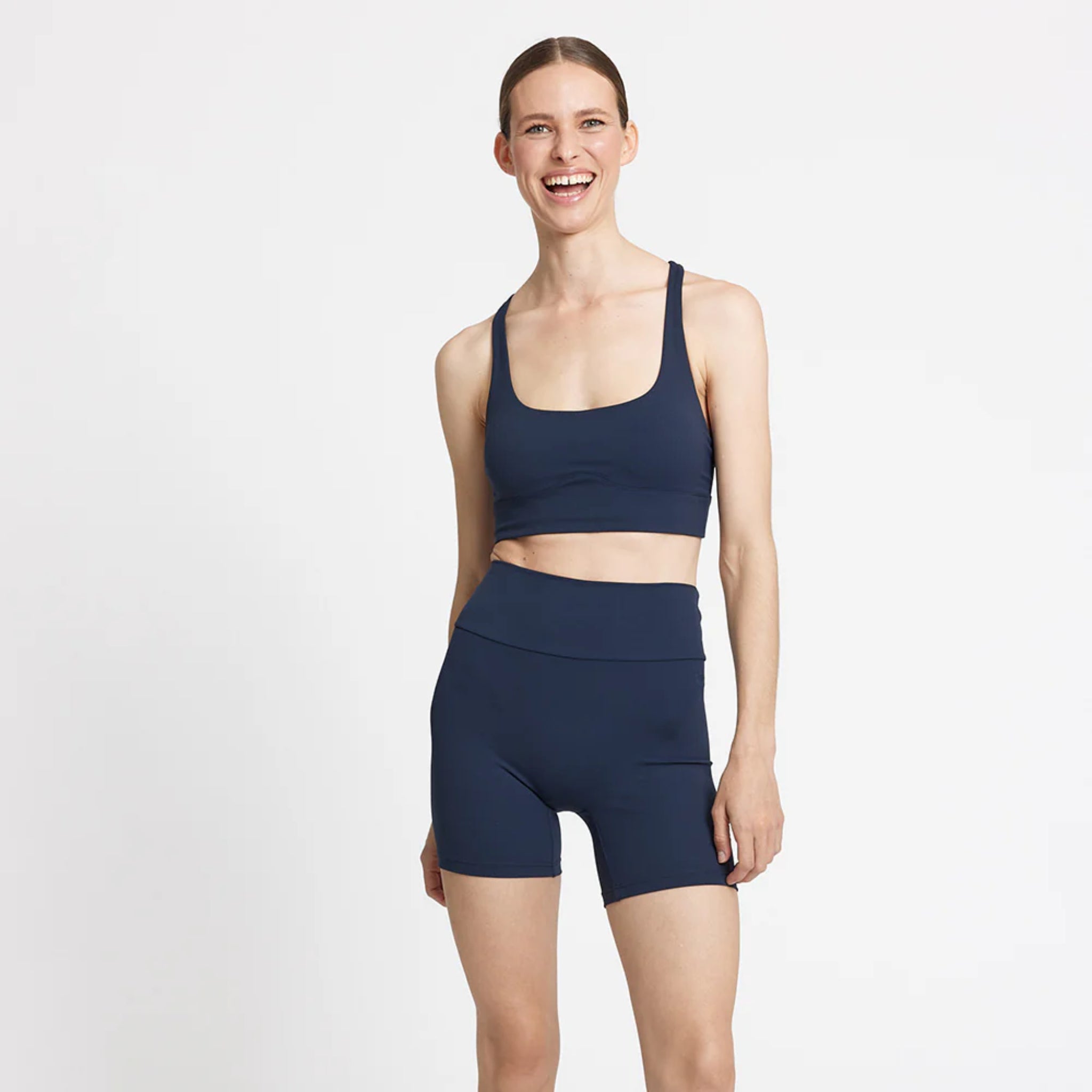 Frau in einem dunkelblauen Sportoberteil und Shorts lächelt vor einem weißen Hintergrund.