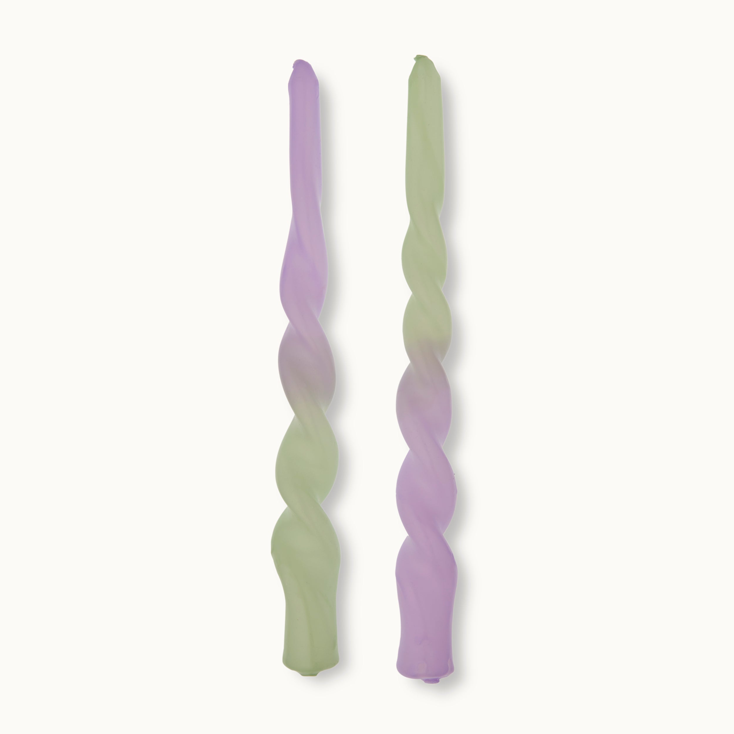 Zwei gedrehte Spitzkerzen, eine in Pastellgrün und eine in Pastellviolett, stehen aufrecht nebeneinander auf einem schlichten weißen Hintergrund.