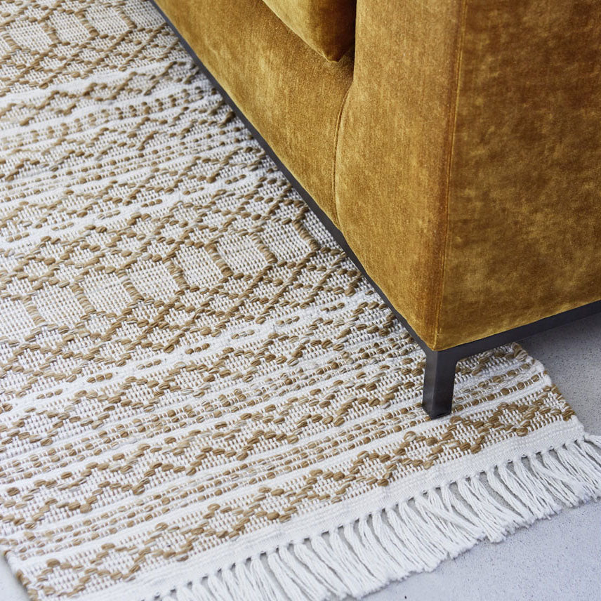 Nahaufnahme eines Sofas aus goldenem Samt mit schwarzen Metallbeinen neben einem gewebten Teppich mit beige-weißem geometrischem Muster und Fransenrand.