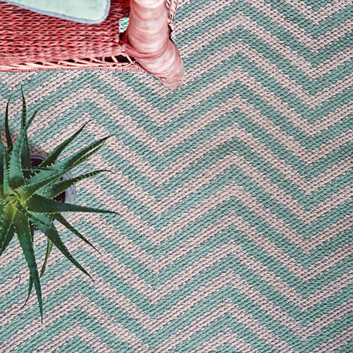 Ein grüner und beiger gewebter Teppich mit Chevron-Muster, auf dem eine grüne Sukkulente und ein Teil eines rosa Korbstuhls in der oberen linken Ecke zu sehen sind.