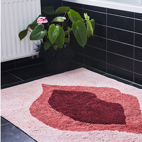 Ein schwarz gefliestes Badezimmer mit einer Badewanne, ein rosa und rot gemusterter Teppich auf dem Boden und eine grüne Pflanze mit rosa Blüten in einem schwarzen Topf neben dem Fenster.