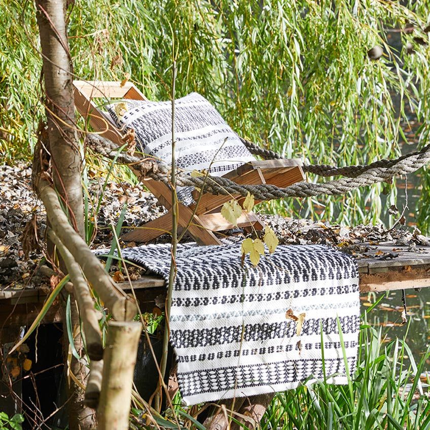 Ein Holzstuhl mit einem gemusterten schwarz-weißen Kissen und einer Decke steht auf einer rustikalen Terrasse in einem üppig begrünten Außenbereich, umgeben von Bäumen und Grünpflanzen.