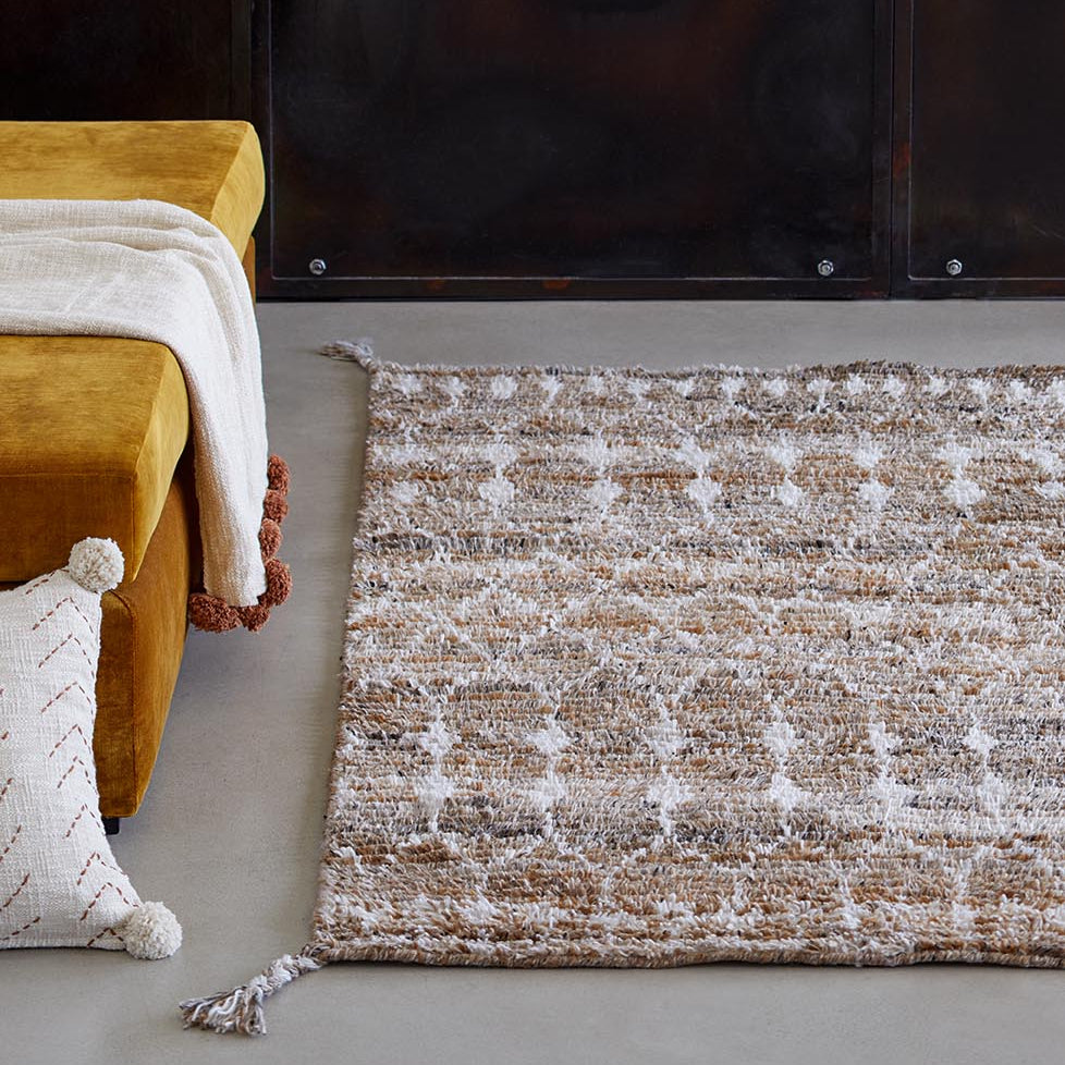 Gelbes Sofa mit weißem Kissen und Decke daneben ein gemusterter Teppich auf grauem Boden.