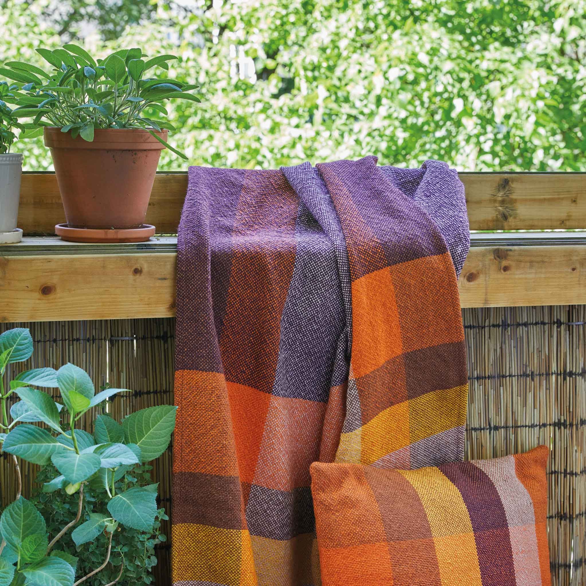 Eine karierte Decke und ein passendes Kissen in Orange, Gelb, Braun und Violett liegen auf einem Balkongeländer mit Topfpflanzen, einem Bambusschirm und Grünpflanzen im Hintergrund.