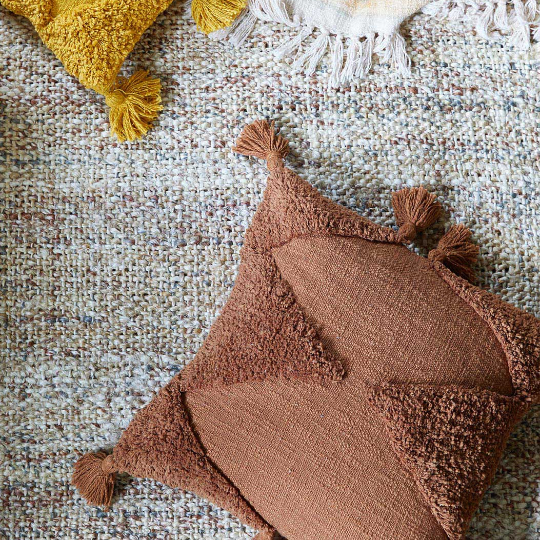 Braunes Kissen mit Quasten auf einem strukturierten, gemusterten Teppich liegend.