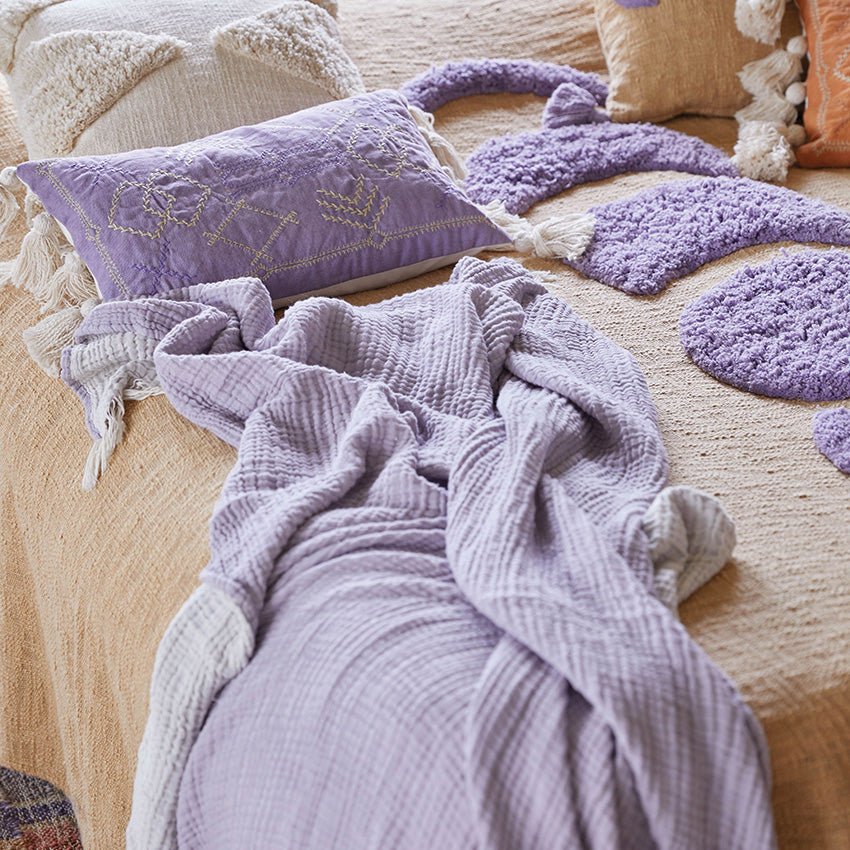 Ein gemütliches, beigefarbenes Bett mit einer lilafarbenen, strukturierten Decke, einem lavendelfarbenen Kissen mit Quasten und einem flauschigen, lilafarbenen Teppich in Katzenform schafft eine weiche und einladende Atmosphäre.