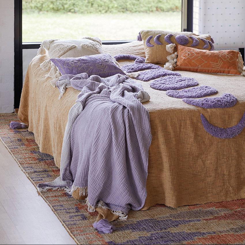 Ein gemütliches Bett mit einer hellbraunen und lila strukturierten Decke, mehreren lila und orangefarbenen Zierkissen und einer lavendelfarbenen Decke steht neben einem großen Fenster mit Blick auf einen grünen Garten. Ein gemusterter Teppich bedeckt den Boden.