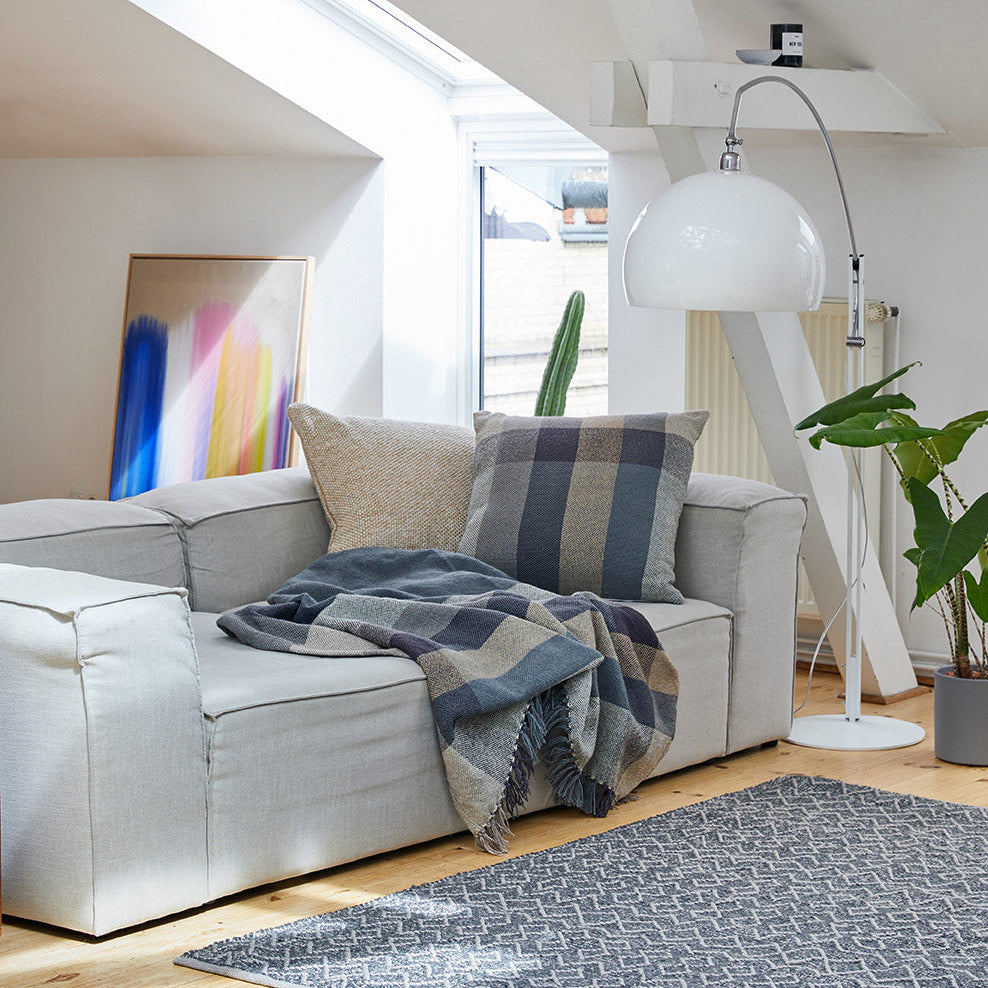 Ein modernes Wohnzimmer mit einem hellgrauen Sofa, karierten und einfarbigen Kissen, einer kuscheligen Decke, einer Stehlampe, einem gemusterten Teppich, einer Topfpflanze und einem farbenfrohen abstrakten Gemälde unter einem Oberlicht.