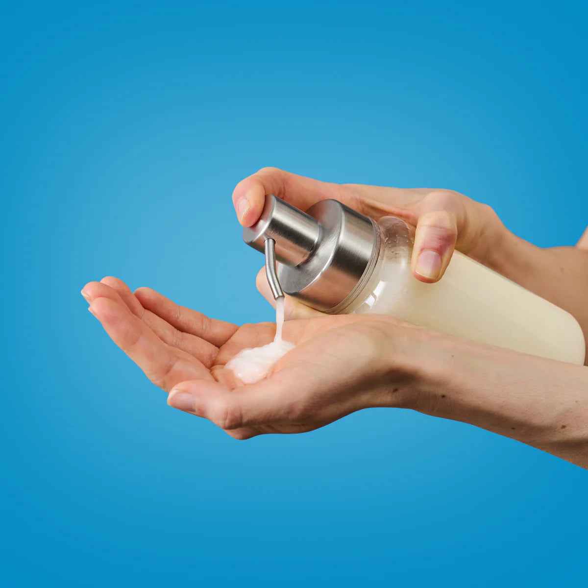 Eine Person pumpt Flüssigseife aus einer durchsichtigen Spenderflasche in ihre offene Handfläche vor einem einfarbig blauen Hintergrund.