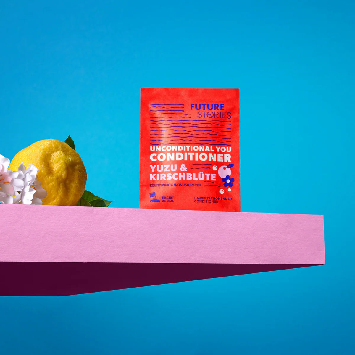 Ein roter Beutel Future Stories Unconditional You Conditioner Yuzu & Kirschblüte steht auf einer rosafarbenen Fläche mit einer gelben Yuzu-Frucht und weißen Blüten, alles vor einem leuchtend blauen Hintergrund.