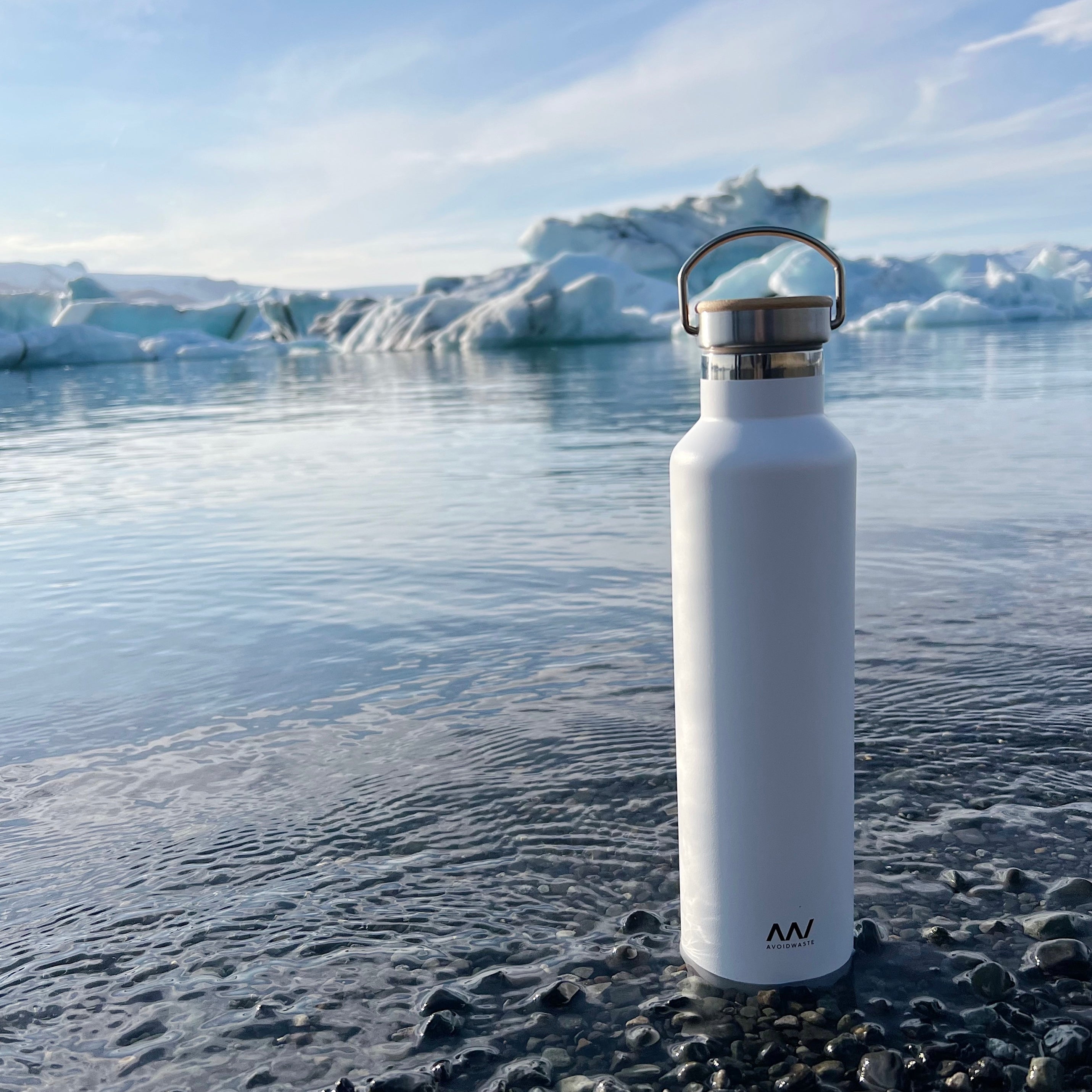 Weiße Wasserflasche steht auf Kieseln am Ufer eines klaren Gewässers mit eisigen Klippen im Hintergrund.