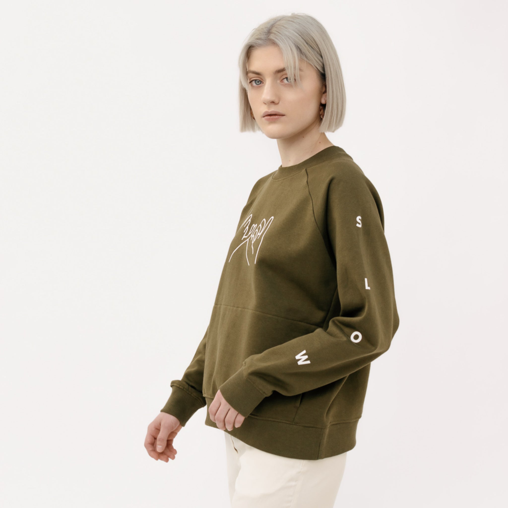 Person mit kurzem, blondem Haar trägt einen khakifarbenen Pullover mit der Aufschrift "SLOW" auf dem Ärmel.