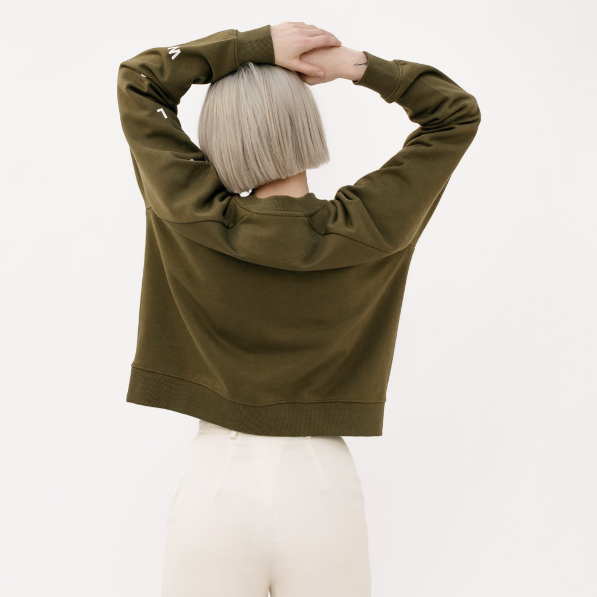 Person mit kurzen blonden Haaren, in einem olivgrünen Pullover, von hinten zu sehen, hebt die Arme über den Kopf.