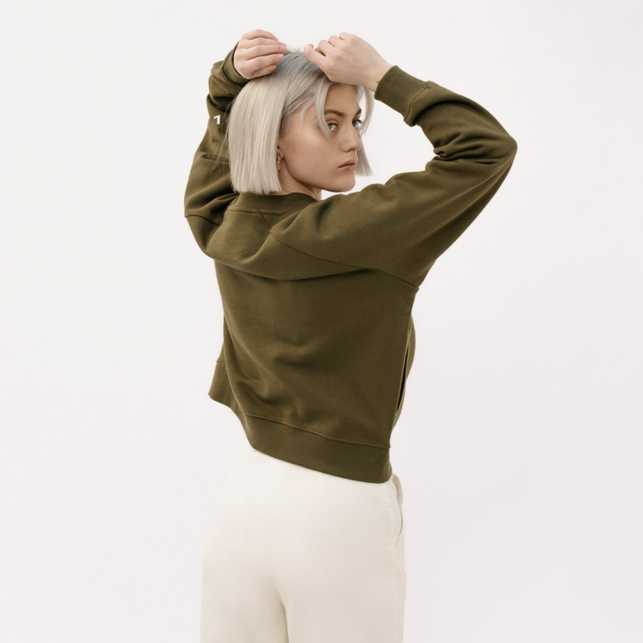 Frau mit blondem Bob trägt einen olivgrünen Pullover und weiße Hose, blickt über die Schulter.