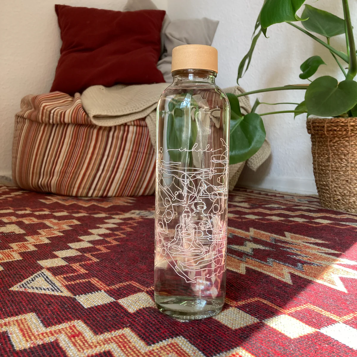 Eine Glasflasche mit Korkverschluss steht auf einem gemusterten Teppich, im Hintergrund sind Kissen und eine Pflanze.