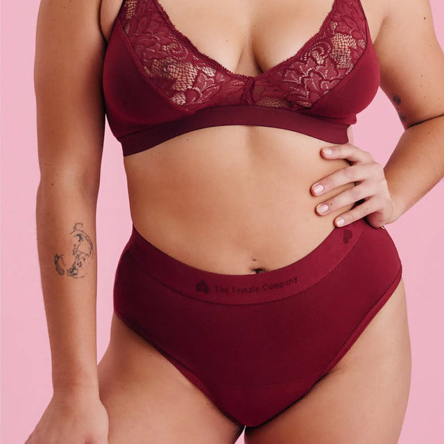 Eine Person, die einen weinroten Spitzen-Bralette und hochtaillierte Unterwäsche trägt, steht vor einem rosafarbenen Hintergrund und zeigt Tätowierungen auf ihrem Oberarm und Oberschenkel.