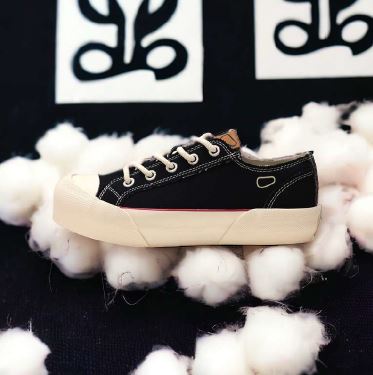 Ein schwarzer Sneaker mit weißen Schnürsenkeln liegt auf weißen Baumwollbällchen vor einem grafischen Hintergrund.