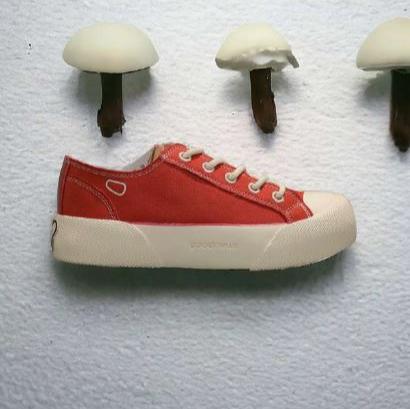 Roter Sneaker an einer weißen Wand, daneben drei Pilze mit braunen Stielen und weißen Kappen.