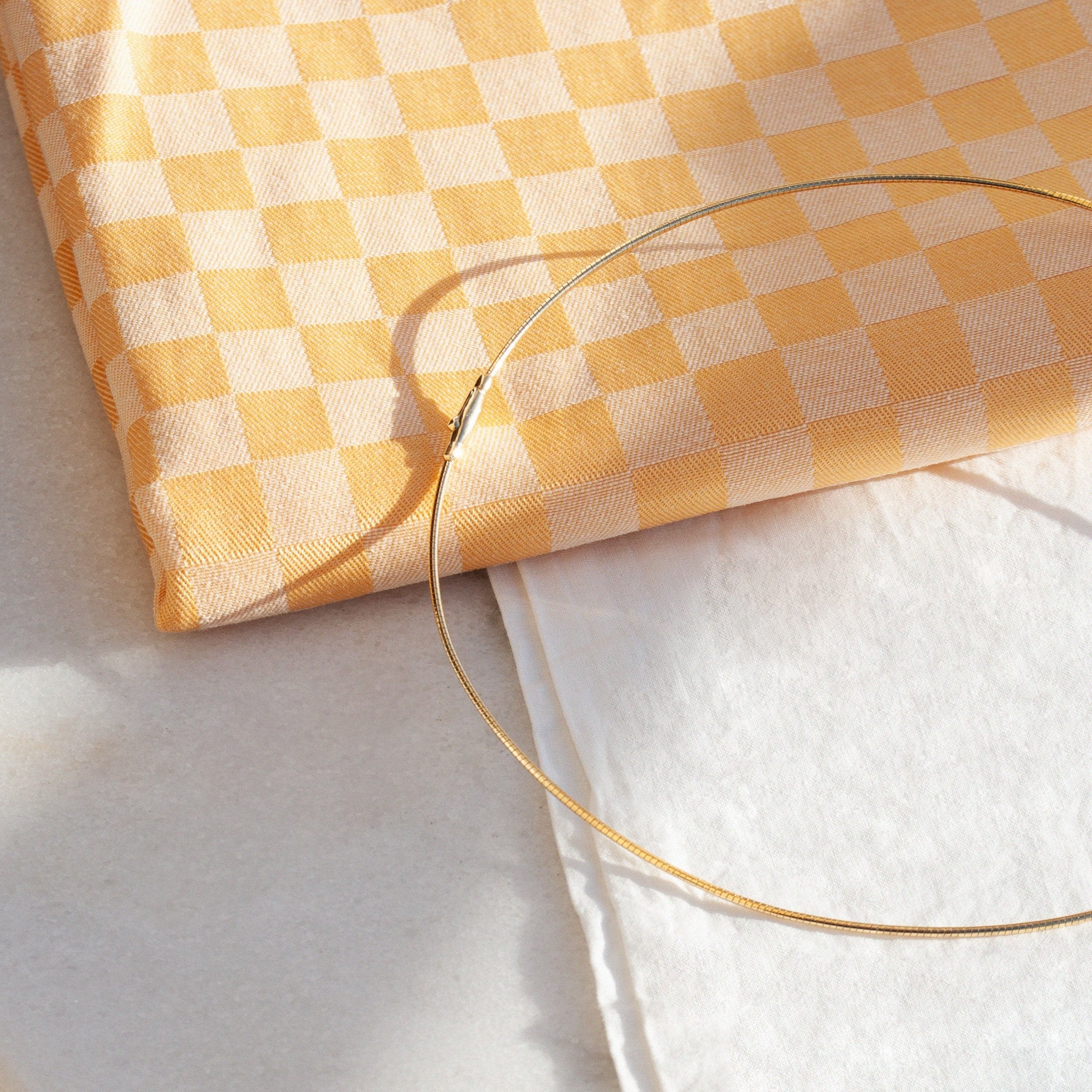 Gelbes karomustertes Tuch neben einem weißen Tuch auf einem Tisch. Ein dünner goldener Ring liegt darauf.