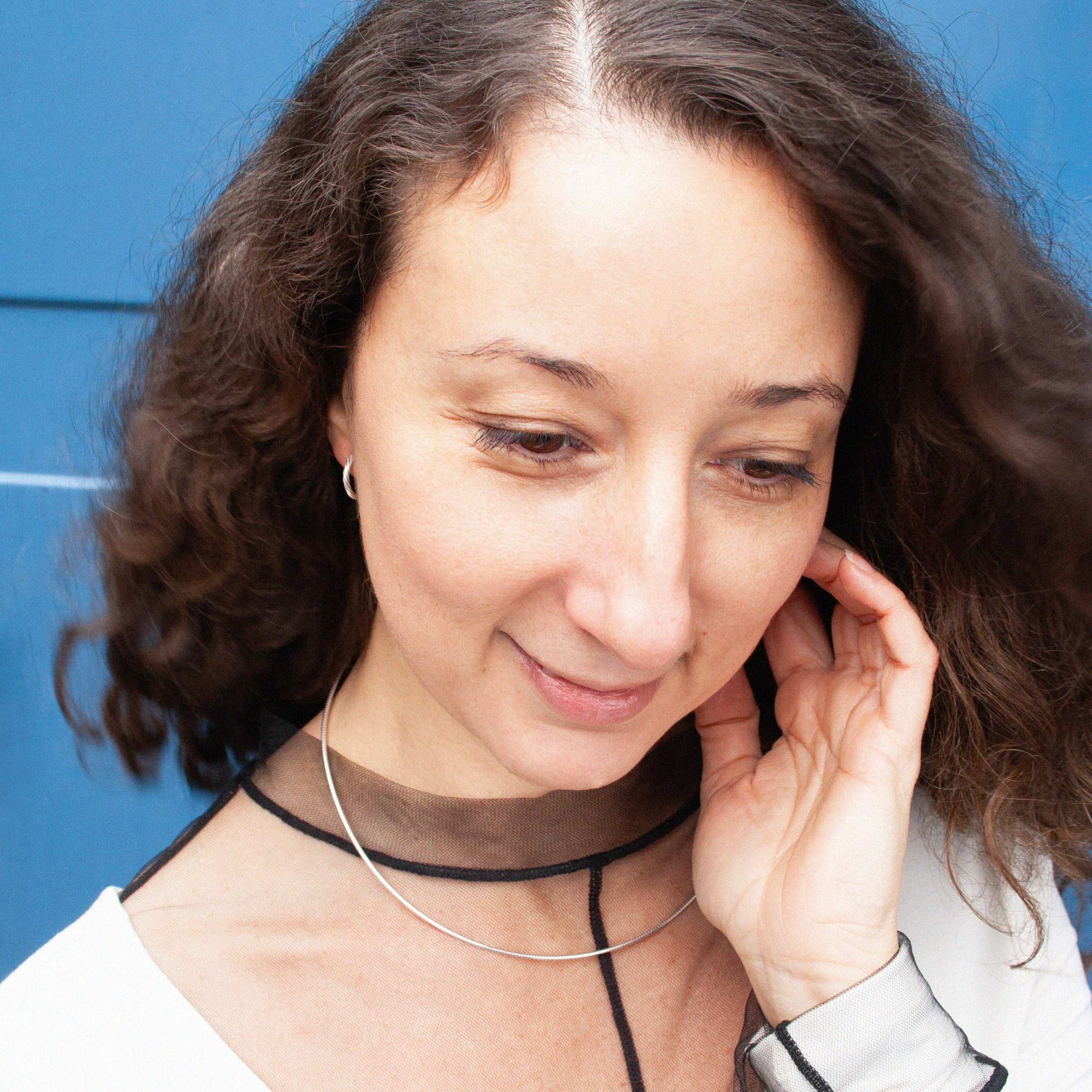 Eine Frau mit lockigem Haar und Ohrringen blickt nach unten, vor einem blauen Hintergrund.